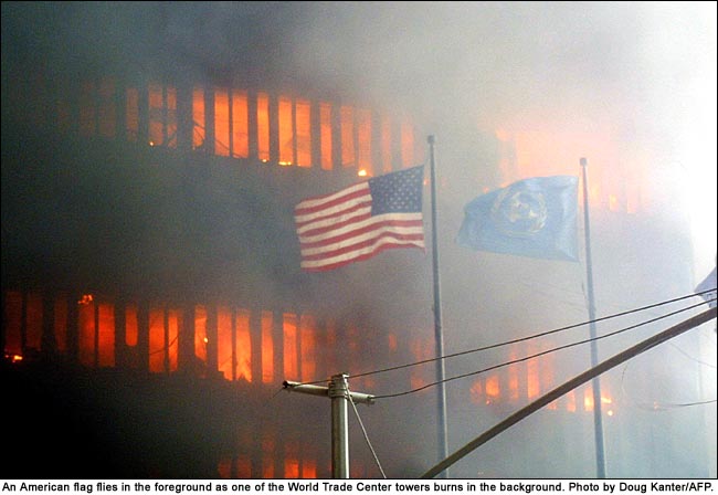 (9/11/01) Flags outside WTC4 not burning, viewed from the east. American flag and UN flag flying in foreground remain unburned while WTC tower burns intensely in background, demonstrating inverse thermal reaction where dielectrics survive.
  <br>
  - Photo by Doug Kanter/AFP